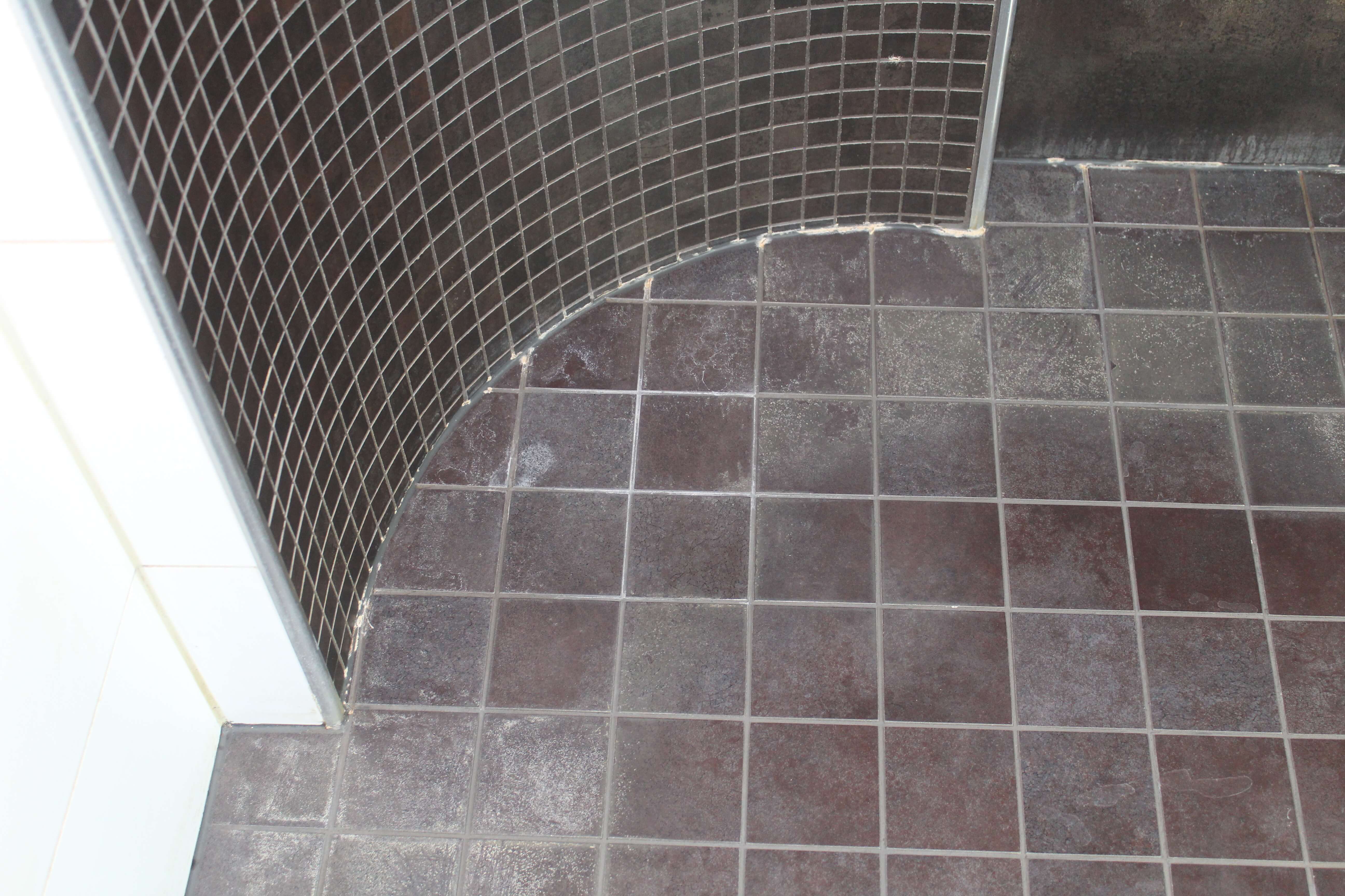 Kylpyhuoneen lattia ja kaareva seinä, joissa on tummat neliömäiset laatat.