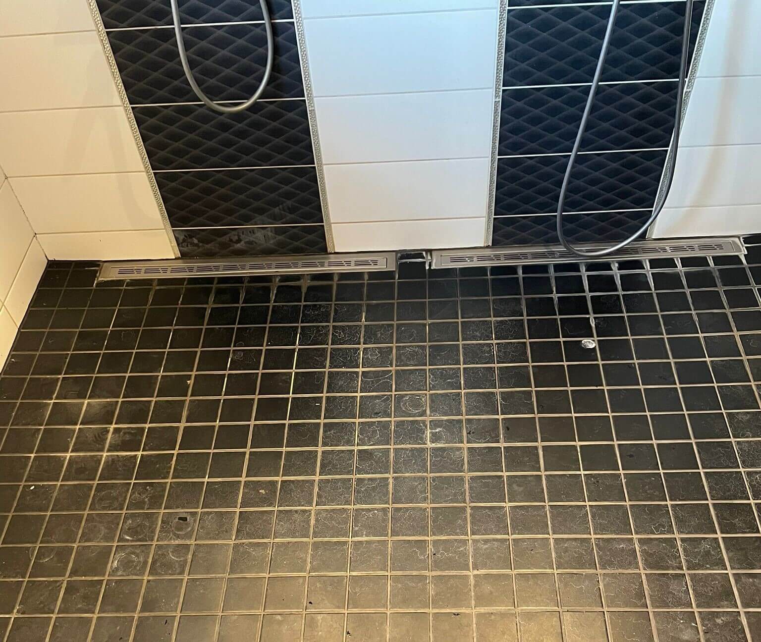 Kaksi mustavalkoista suihkuseinää huoneen tummalla ruutulattialla, jossa on kaksi pitkä ilmastointiritilää seinän vieressä.