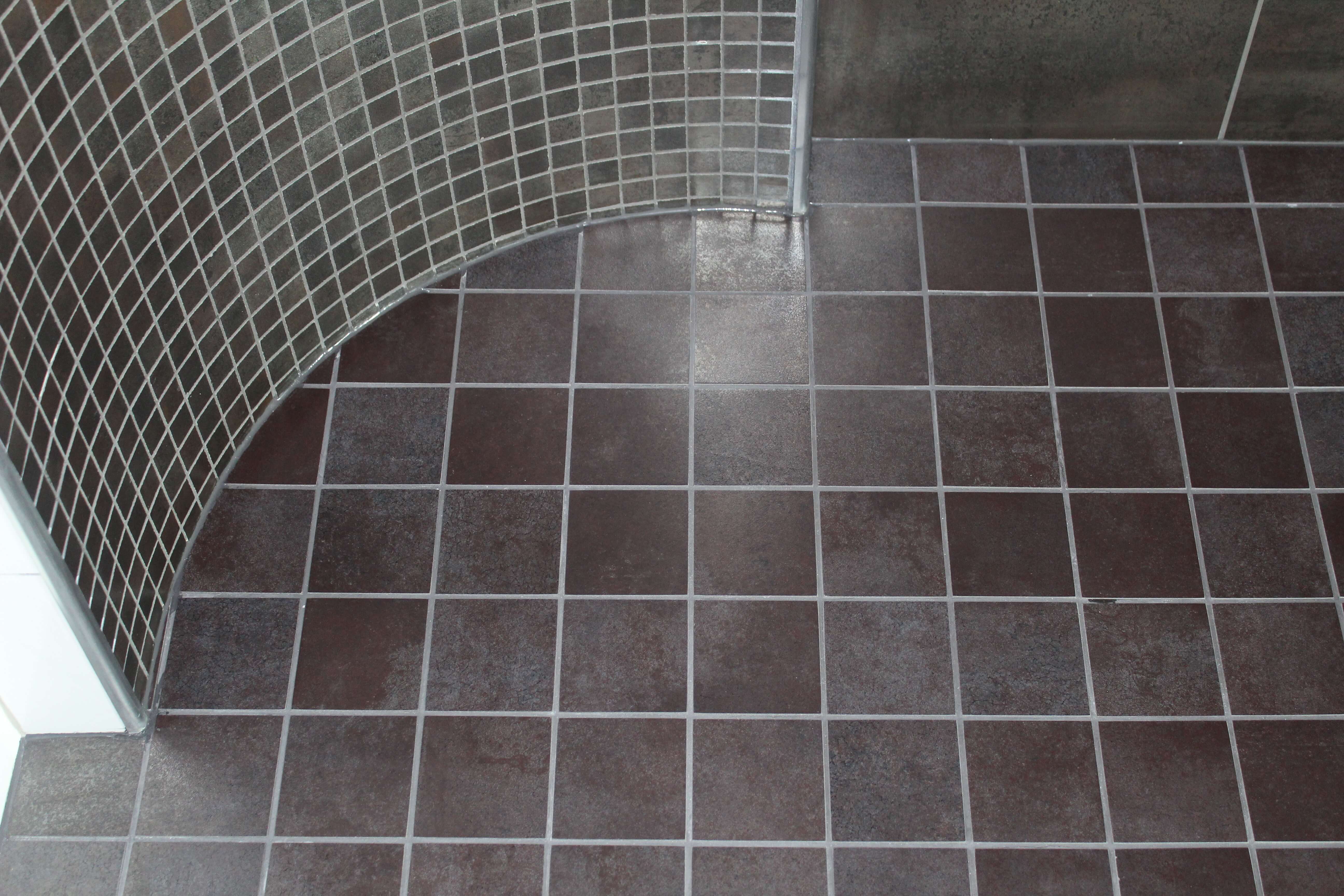 Suihkutilan lattia ja seinät, joissa tumma harmaat neliönmuotoiset laatat, joissa vaaleat saumasaumat ja kaareva suihkualue.