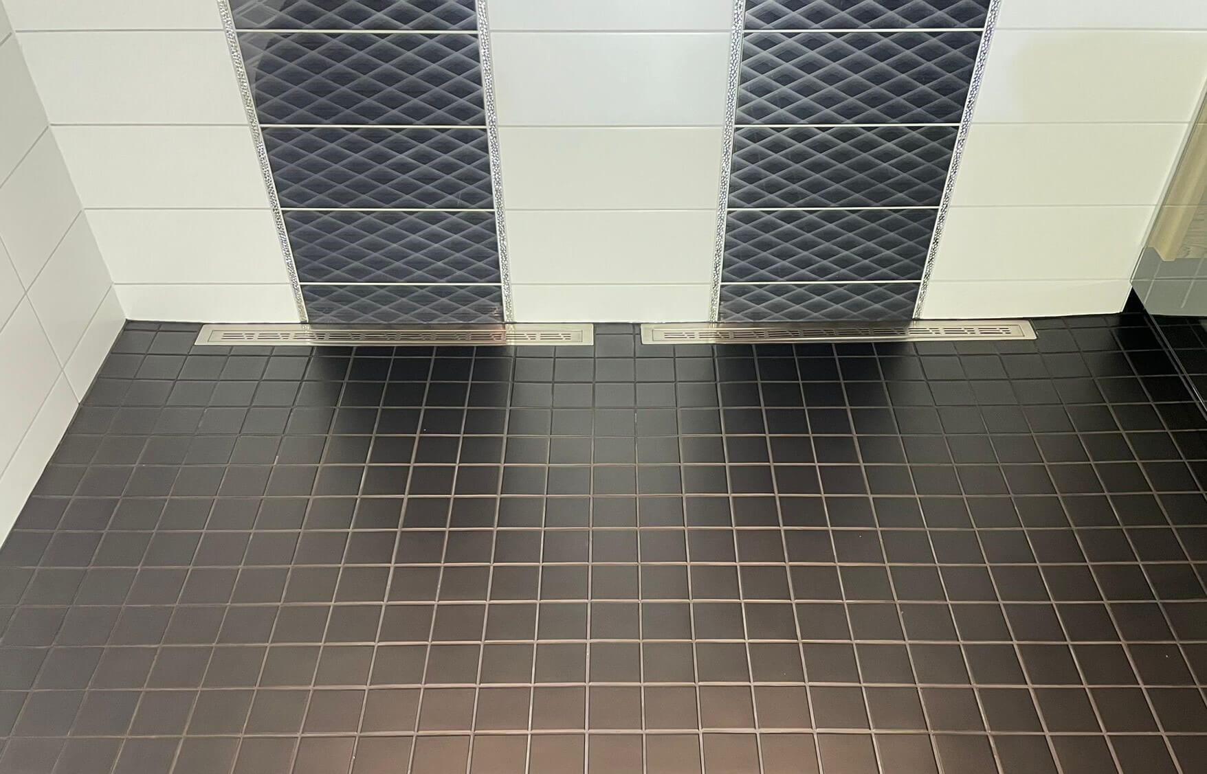 Kylpyhuoneen seinä, jossa on vaaleita ja mustia ruudullisia laattoja ja kaksi lattiakaivoa.